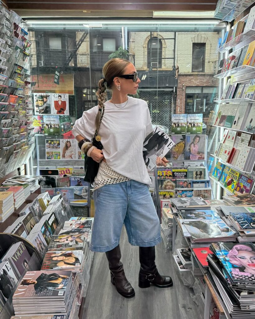 Mulher jovem, magra, alta e de pele clara. Ela está em uma banca de revistas. Ela usa look com bermuda jeans, camiseta branca, lenço na cintura e bota de cano médio.