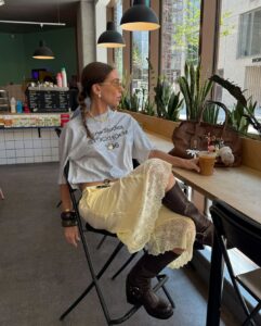 Mulher usando look com bota no verão. Ela veste camiseta de manga curta, saia midi de cetim e rendas, bota de cano médio marrom e usa acessórios como pulseiras, colar, brinco e óculos de grau.