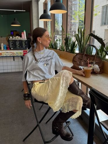 Mulher usando look com bota no verão. Ela veste camiseta de manga curta, saia midi de cetim e rendas, bota de cano médio marrom e usa acessórios como pulseiras, colar, brinco e óculos de grau.