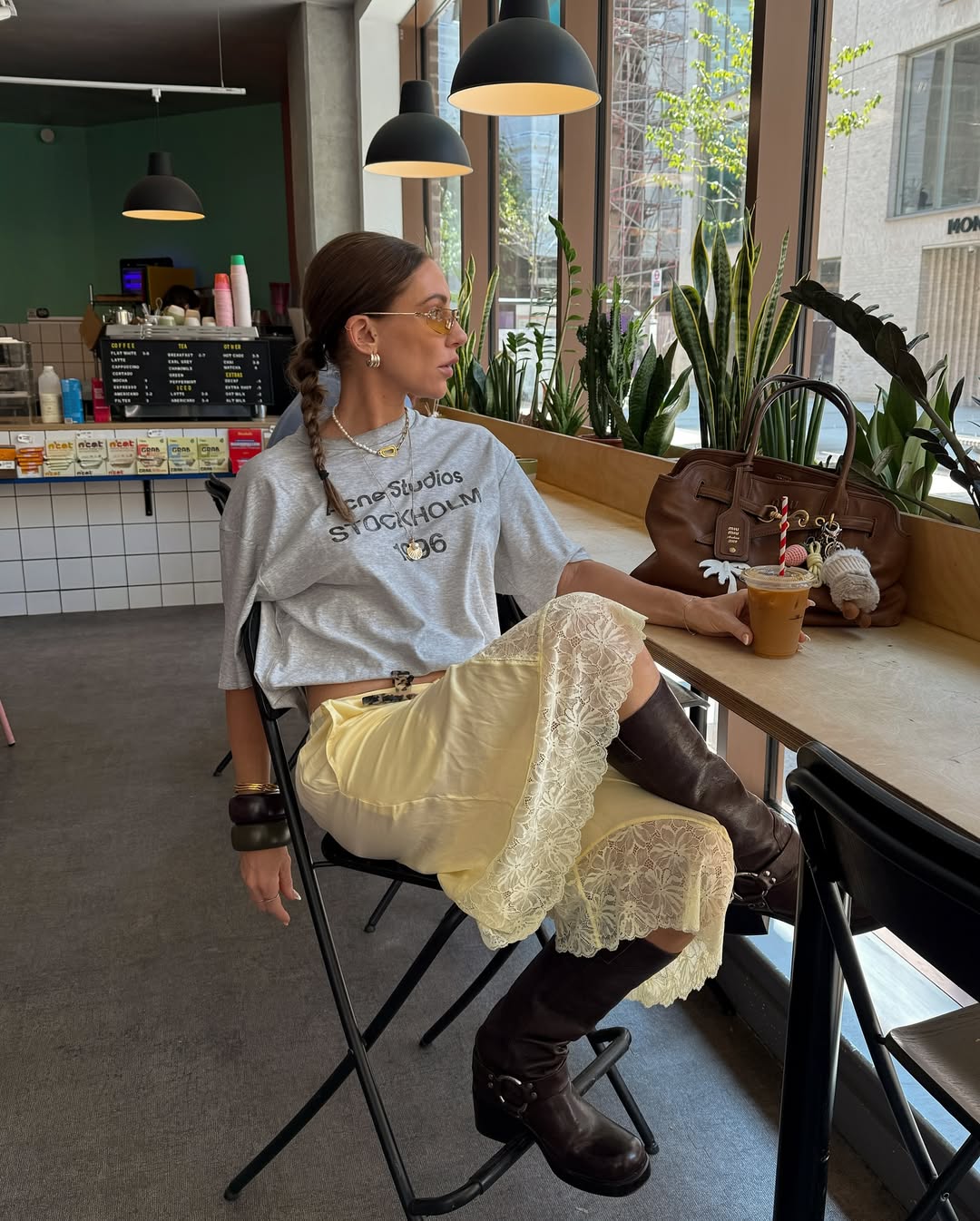 Mulher usando look com bota no verão. Ela veste camiseta de manga curta, saia midi de cetim e rendas, bota de cano médio marrom e usa acessórios como pulseiras, colar, brinco e óculos de grau.