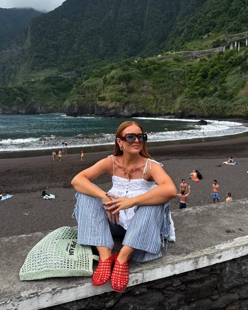 Mulher jovem e magra com pele clara. Ela usa bata branca, calça listrada, bolsa de crochê verde e sapatilha telada vermelha