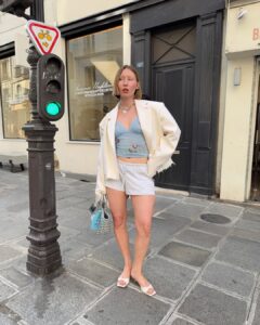 Mulher jovem e magra de pele clara e cabelo loiro curto. Ela usa blusa, bermuda, blazer, tamanco e bolsa.