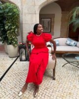 Mulher jovem de pele negra e cabelos escuros escorada em um sofá. Ela usa look de natal com conjunto vermelho e sapatilha dourada