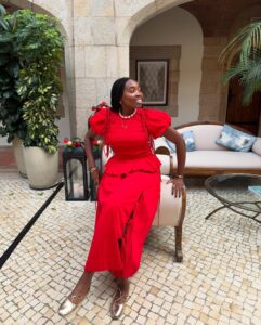 Mulher jovem de pele negra e cabelos escuros escorada em um sofá. Ela usa look de natal com conjunto vermelho e sapatilha dourada