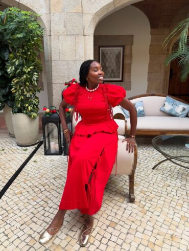 Mulher jovem de pele negra e cabelos escuros escorada em um sofá. Ela usa look de natal com conjunto vermelho e sapatilha dourada