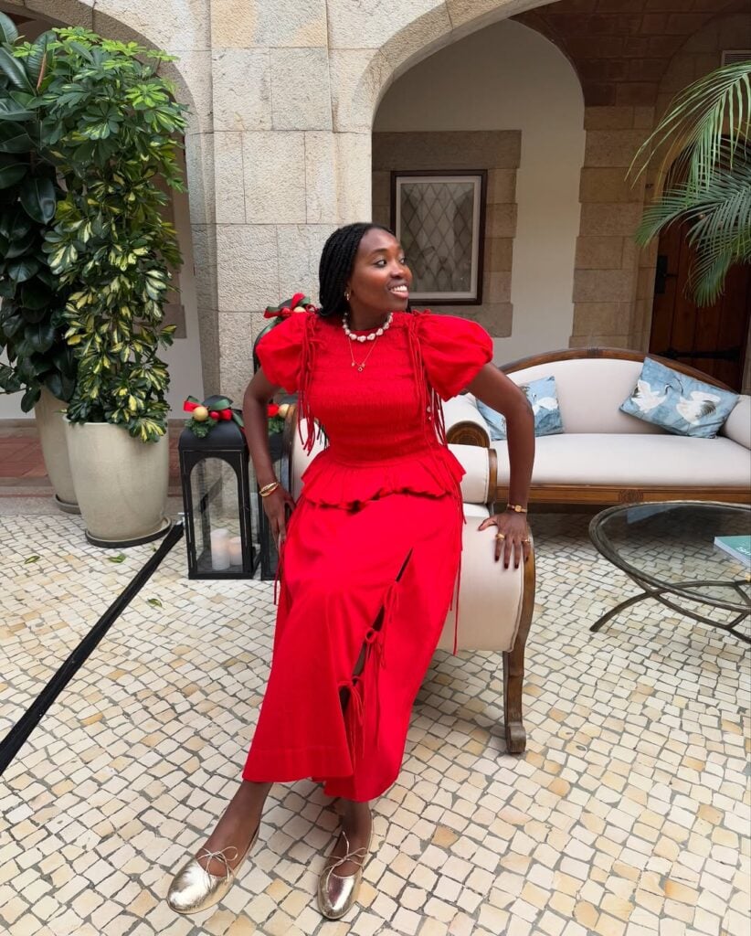 Mulher jovem de pele negra e cabelos escuros escorada em um sofá. Ela usa look de natal com conjunto vermelho e sapatilha dourada
