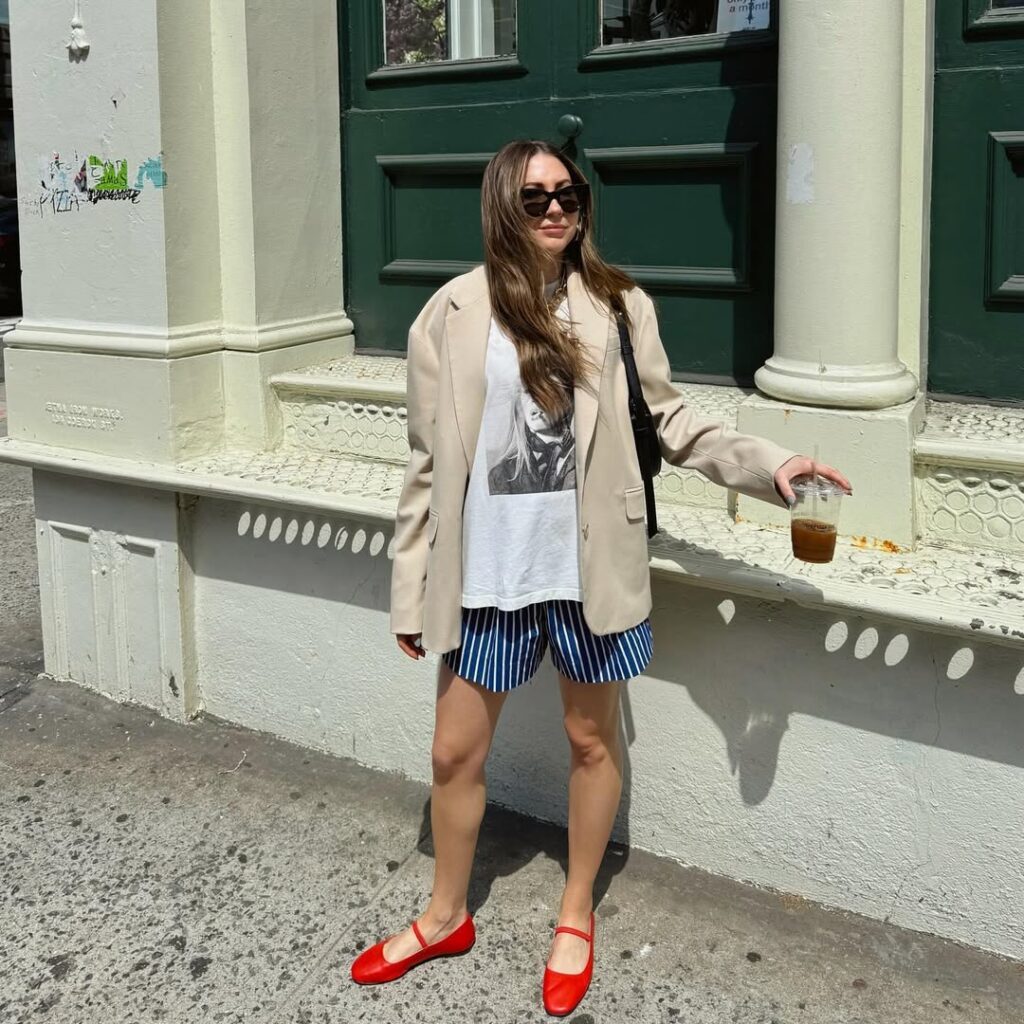 Mulher jovem de pele clara, cabelos longos e castanhos e média estatura. Ela está na frente de uma construção e usa loook de Natal com blazer, camiseta, bermuda e sapatilha