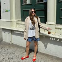 Mulher jovem de pele clara, cabelos longos e castanhos e média estatura. Ela está na frente de uma construção e usa look de Natal com blazer, camiseta, bermuda e sapatilha