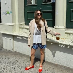 Mulher jovem de pele clara, cabelos longos e castanhos e média estatura. Ela está na frente de uma construção e usa look de Natal com blazer, camiseta, bermuda e sapatilha