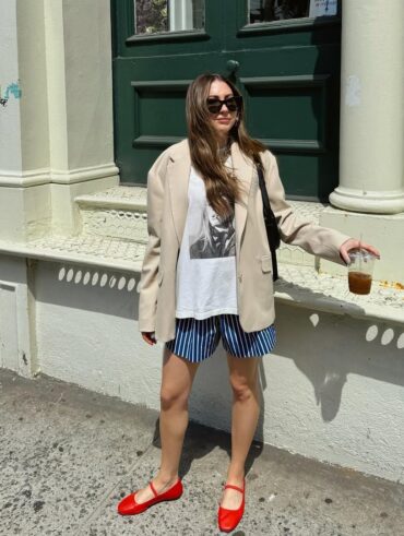 Mulher jovem de pele clara, cabelos longos e castanhos e média estatura. Ela está na frente de uma construção e usa look de Natal com blazer, camiseta, bermuda e sapatilha