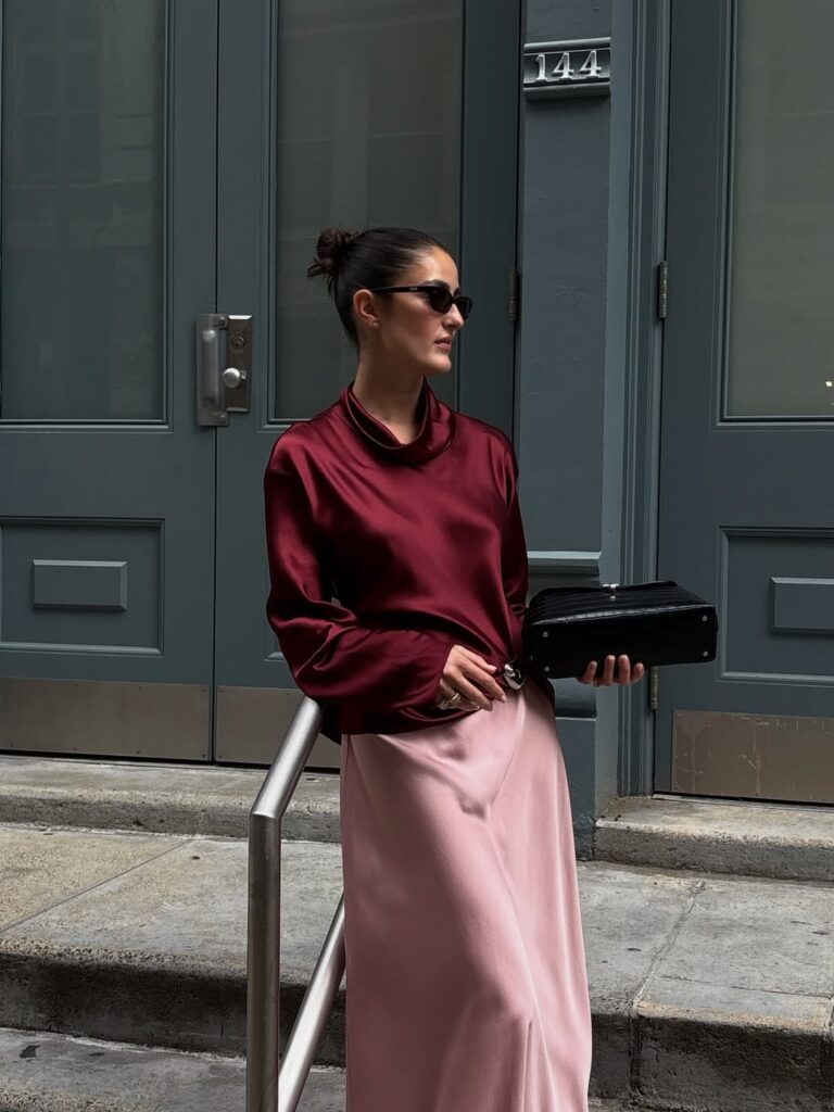 Mulher jovem com camisa de manga longa bordô, saia longa rosa claro, e óculos de sol preto.