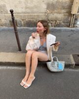 Mulher jovem e magra de pele clara. Ela está sentada em uma calçada e usa tamanco branco, blazer creme, shorts e blusa.