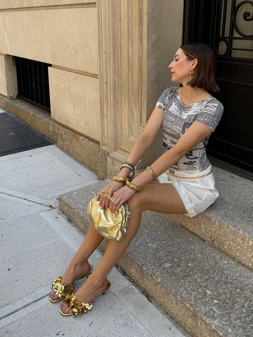 Mulher jovem e magra. Ela usa camiseta, saia, sandália de salto alto, bolsa de mão e pulseiras