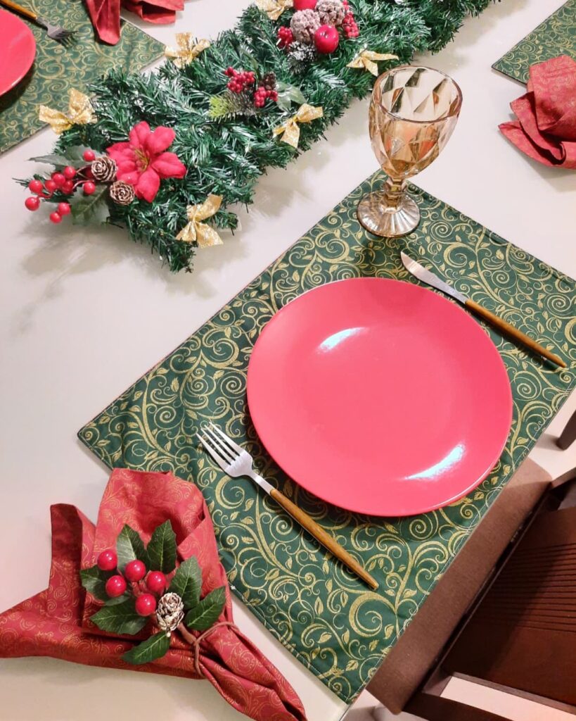 Mesa branca com jogo americano verde e dourado, prato vermelho e guardanapo com azevinho