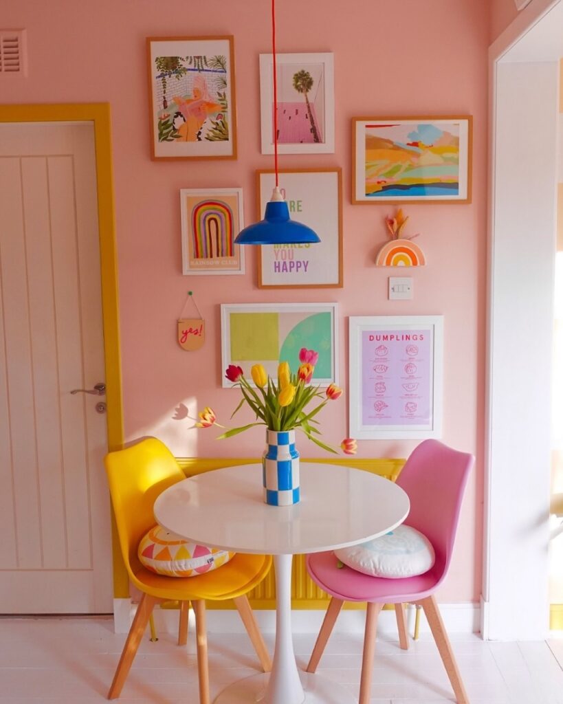 Ambiente com parede rosa e quadros decorativos, mesa branca redonda e cadeiras amarela e rosa