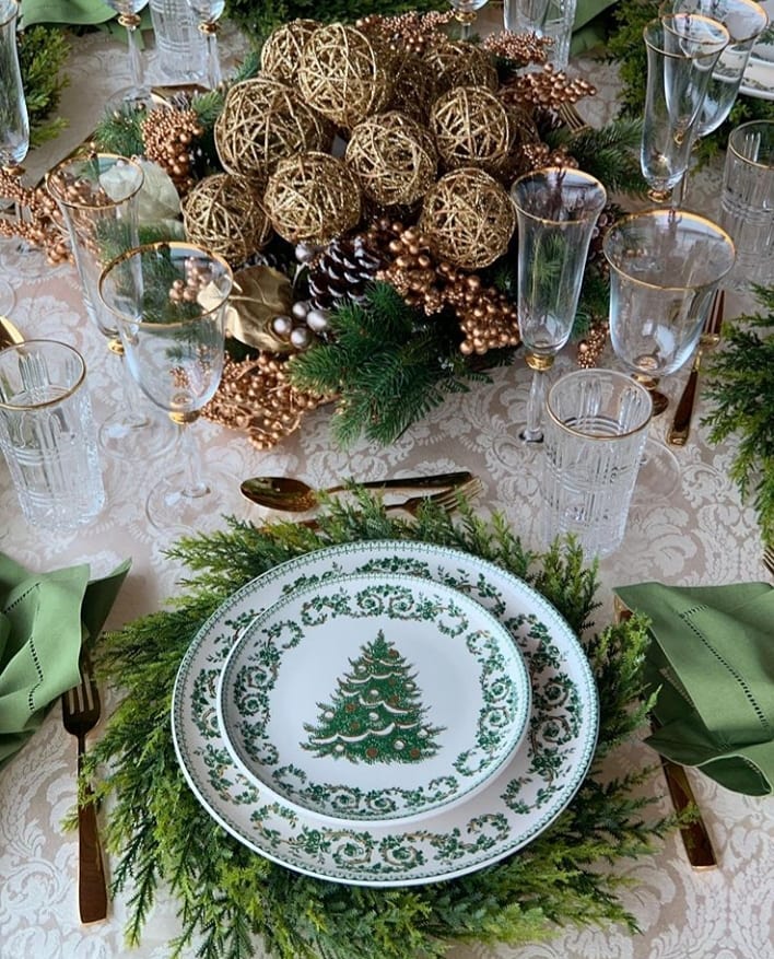 Mesa posta de Natal com pratos brancos decorados com pinheirinhos, taças com bordas douradas e centro de mesa com pinhas e bolas douradas