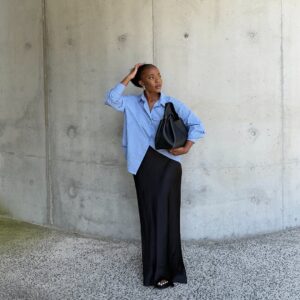Mulher jovem e magra de pele negra. Ela veste camisa social azul, saia longa acetinada e chinelo