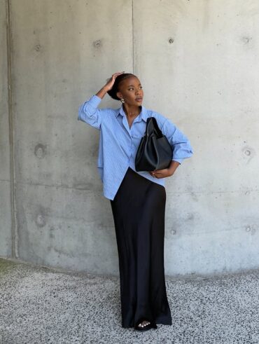 Mulher jovem e magra de pele negra. Ela veste camisa social azul, saia longa acetinada e chinelo