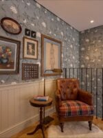 Ambiente com papel de parede floral, quadros e poltrona marrom em couro e assento xadrez