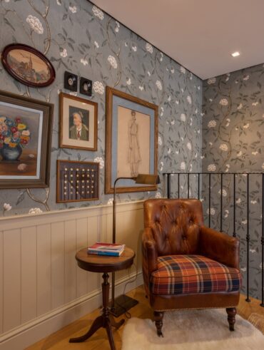 Ambiente com papel de parede floral, quadros e poltrona marrom em couro e assento xadrez