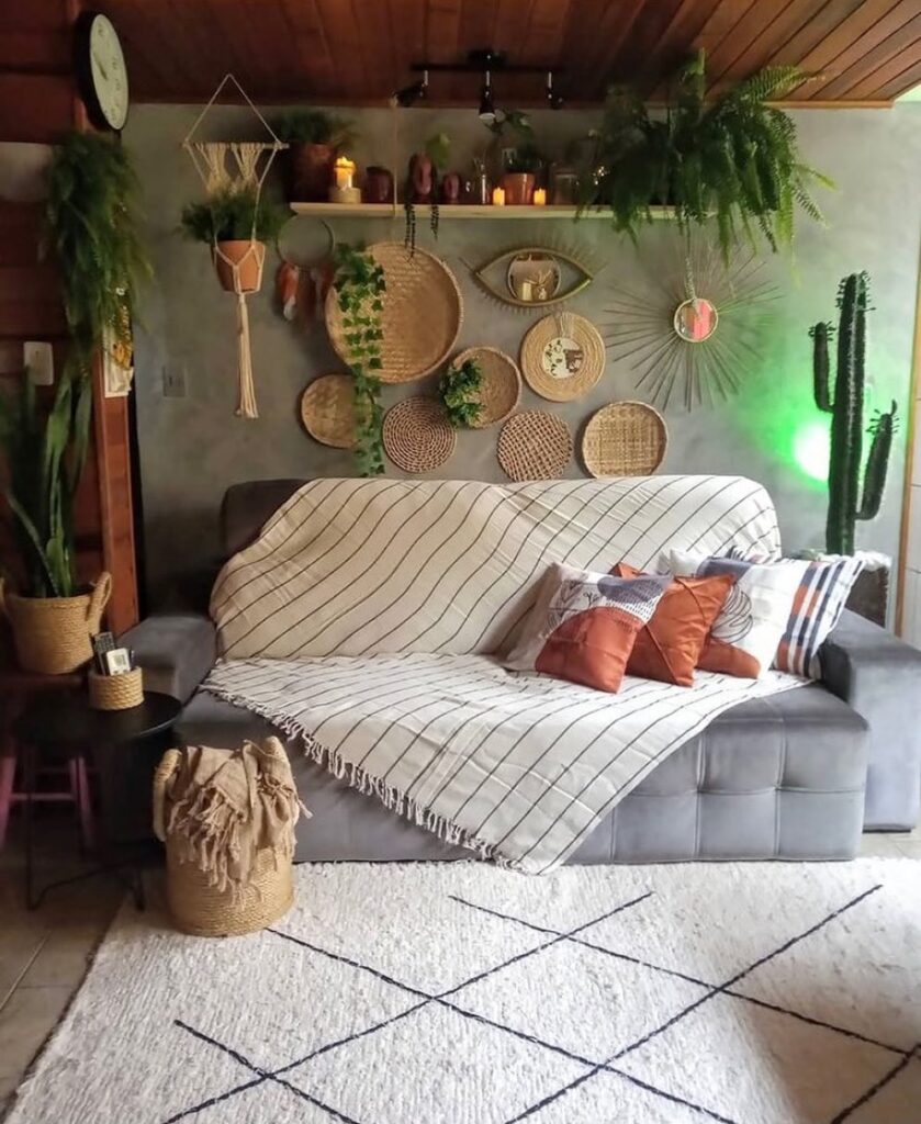 Sala no estilo boho, com sofá cinza em veludo, manta clara com almofadas terrosas, itens de palha na parede e tapete bege com linhas pretas