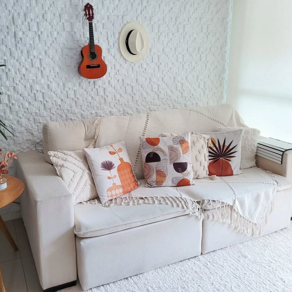 Salla com paredes e sofá branco, almofadas com estampas, violão e chapéu pendurado na parede