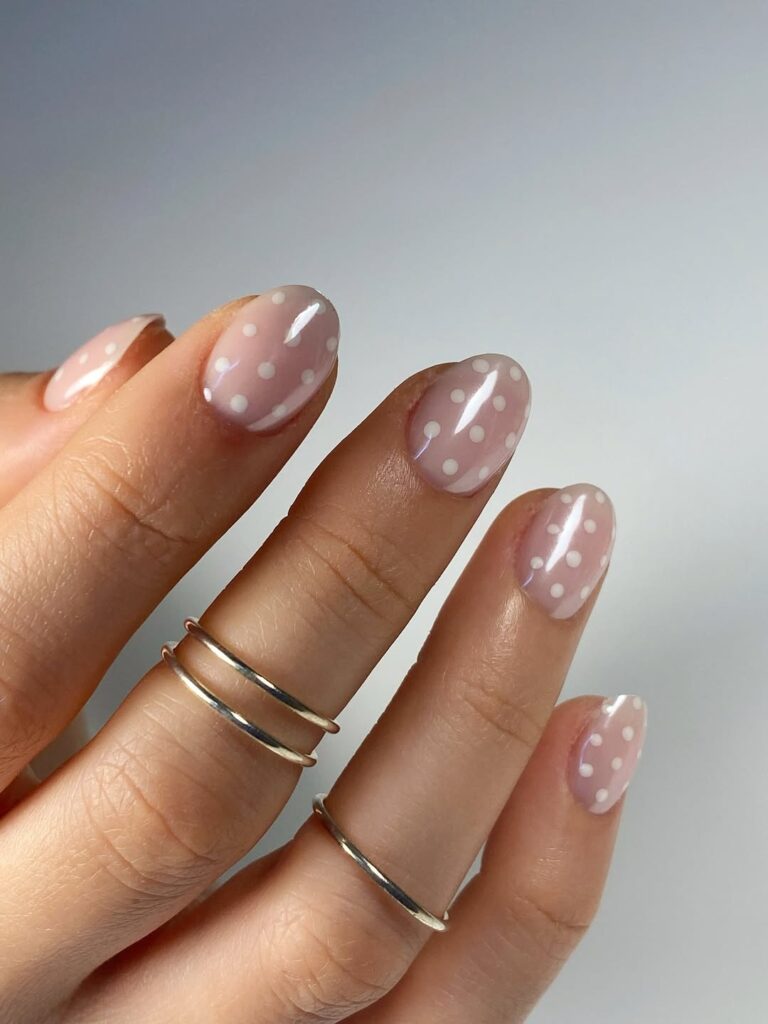 Mãos de pele clara, com anéis em prata e unhas nude rosadas com poá