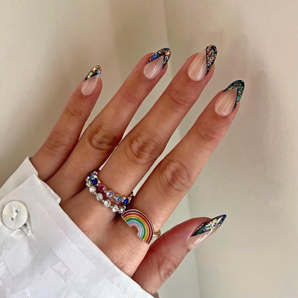 Mão de pele clara, com anéis coloridos e unha amendoada com decorações holográficas 