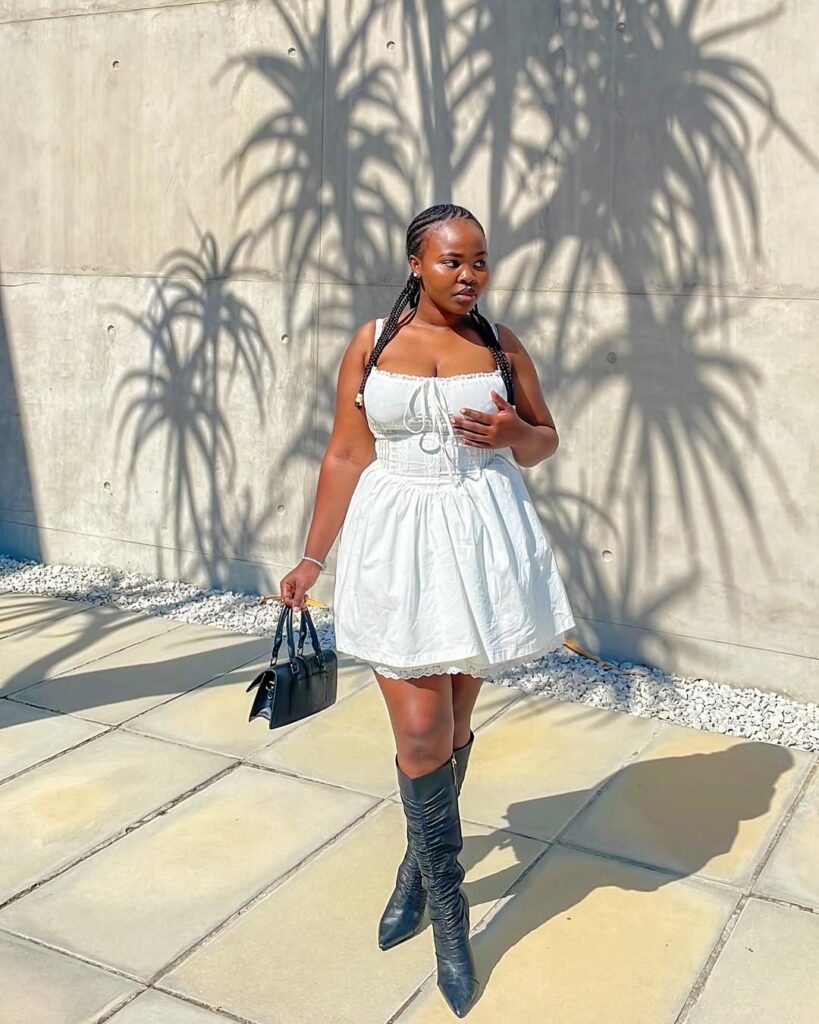 Mulher jovem com vestido branco, bota de cano alto preta e bolsa preta pequena