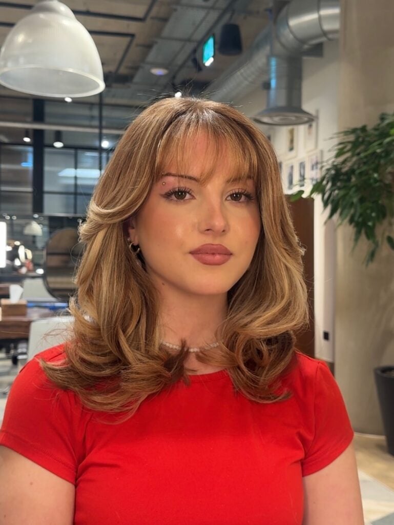 Mulher de pele clara, com blusa vermelha, e cabelo loiro médio com wispy bangs 