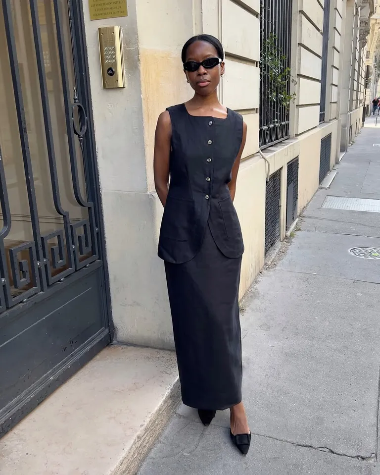 Mulher jovem e magra com look all black de colete e saia longa e sapato de bico fino