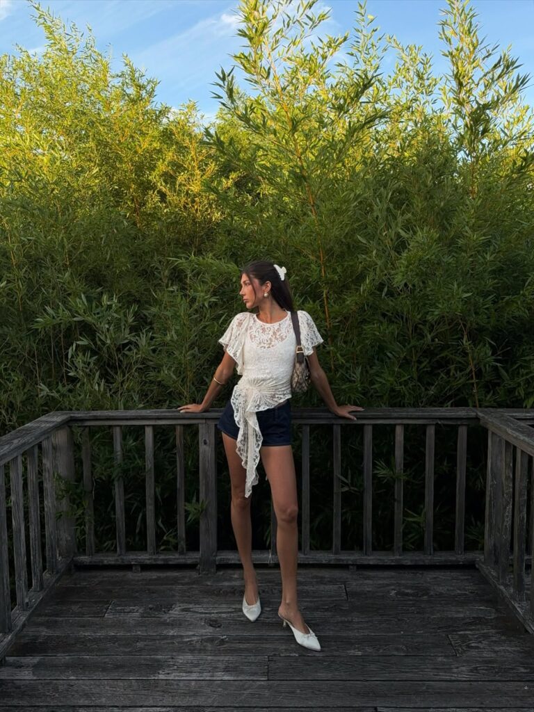 Mulher jovem, magra e de pele clara. Ela usa blusa com detalhes em renda, shorts jeans azul marinho e salto de bico fino.
