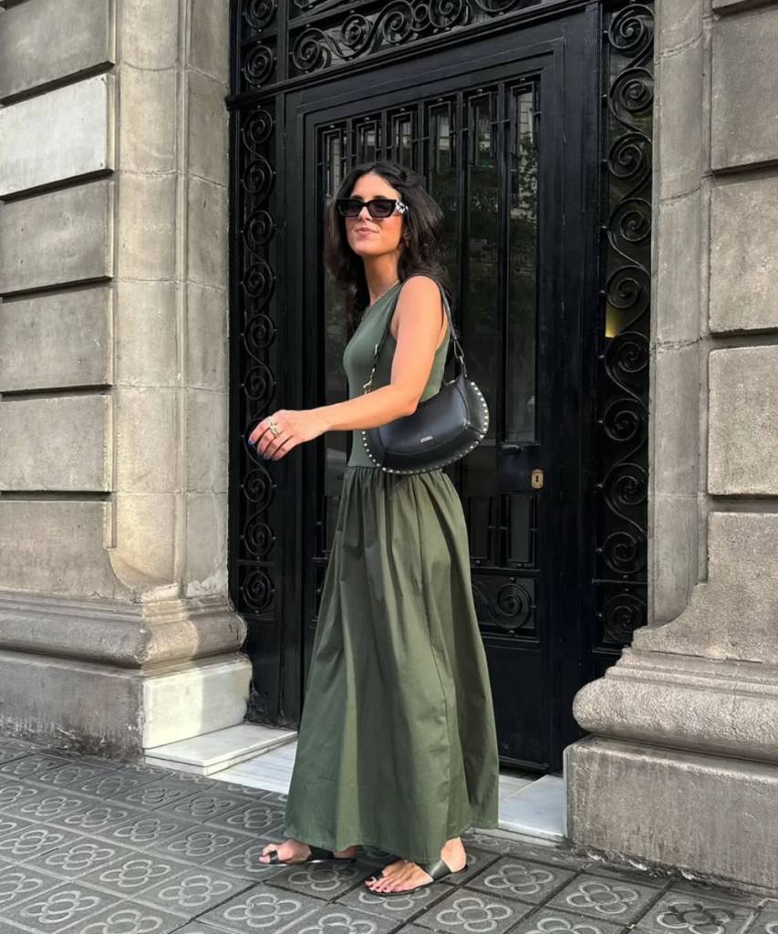 Mulher jovem e magra d epele clara na frente de uma porta. Ela usa vestido verde oliva, sandália preta, bolsa preta e óculos de sol