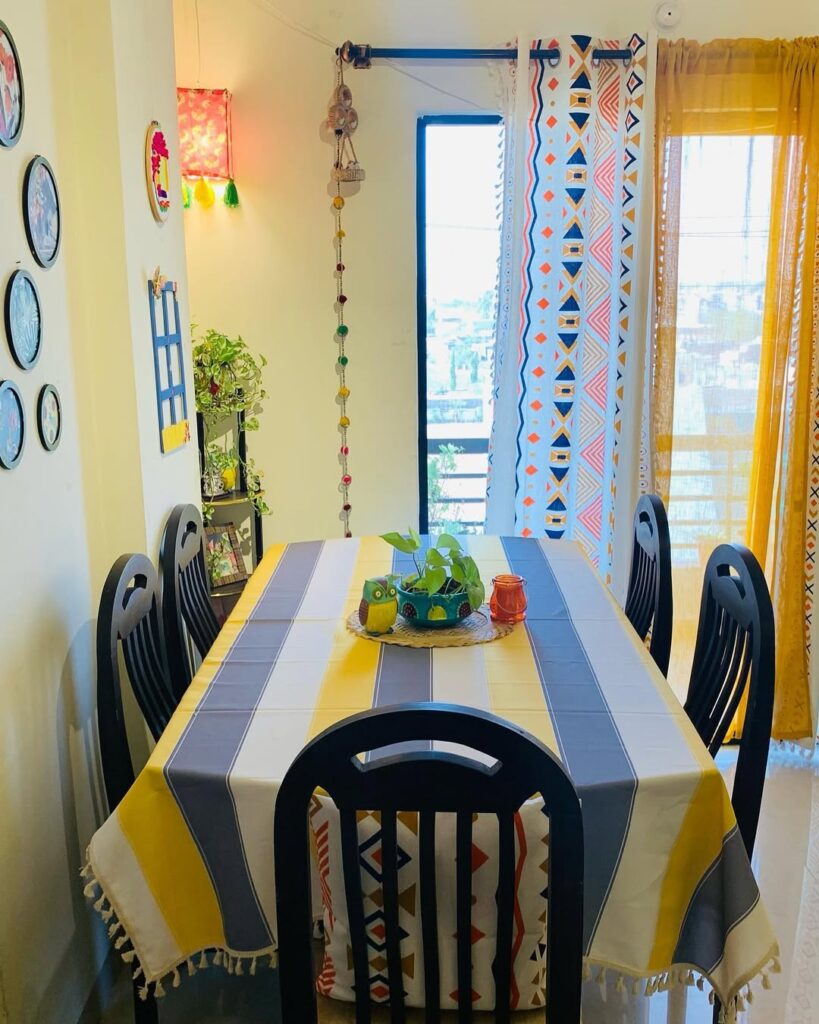 Mesa com toalha listrada, cadeiras azul-marinho e cortinas amarela e colorida