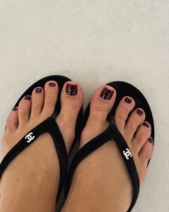 Pés de pele clara, com unhas escuras. Ela está usando um chinelo preto da Chanel