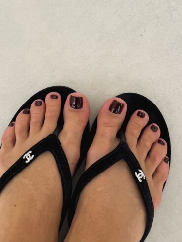 Pés de pele clara, com unhas escuras. Ela está usando um chinelo preto da Chanel