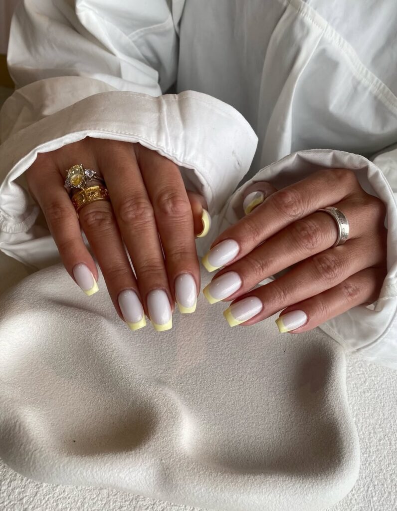 Mãos de pele clara, com anéis em dourado e unha branca com francesinha amarelo-manteiga 