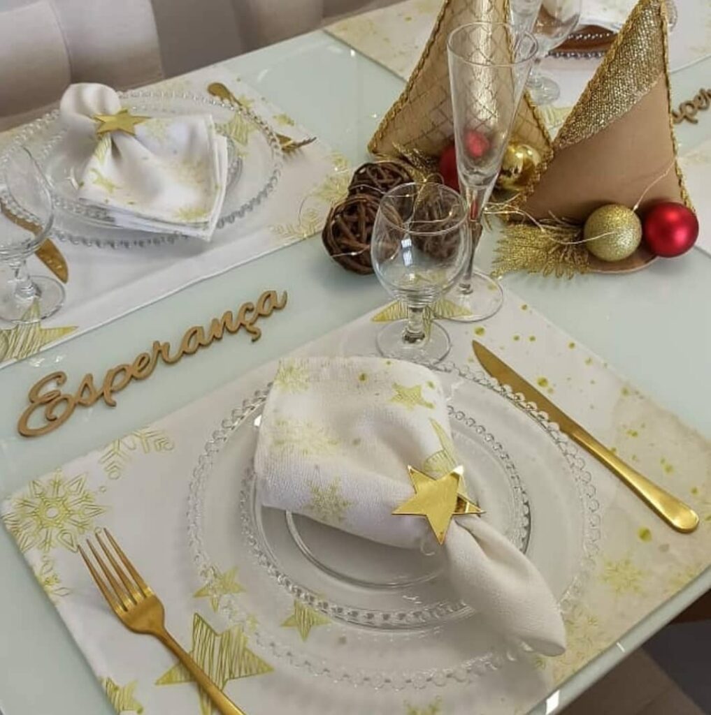 Mesa de Ano-Novo com jogo americano branco com estrelas douradas, talheres dourados, e enfeites natalinos
