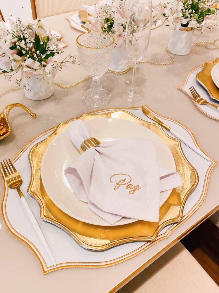 Ideia de mesa posta de Ano-Novo com jogo americano branco, prato e detalhes em dourado, e pequenos arranjos de flores