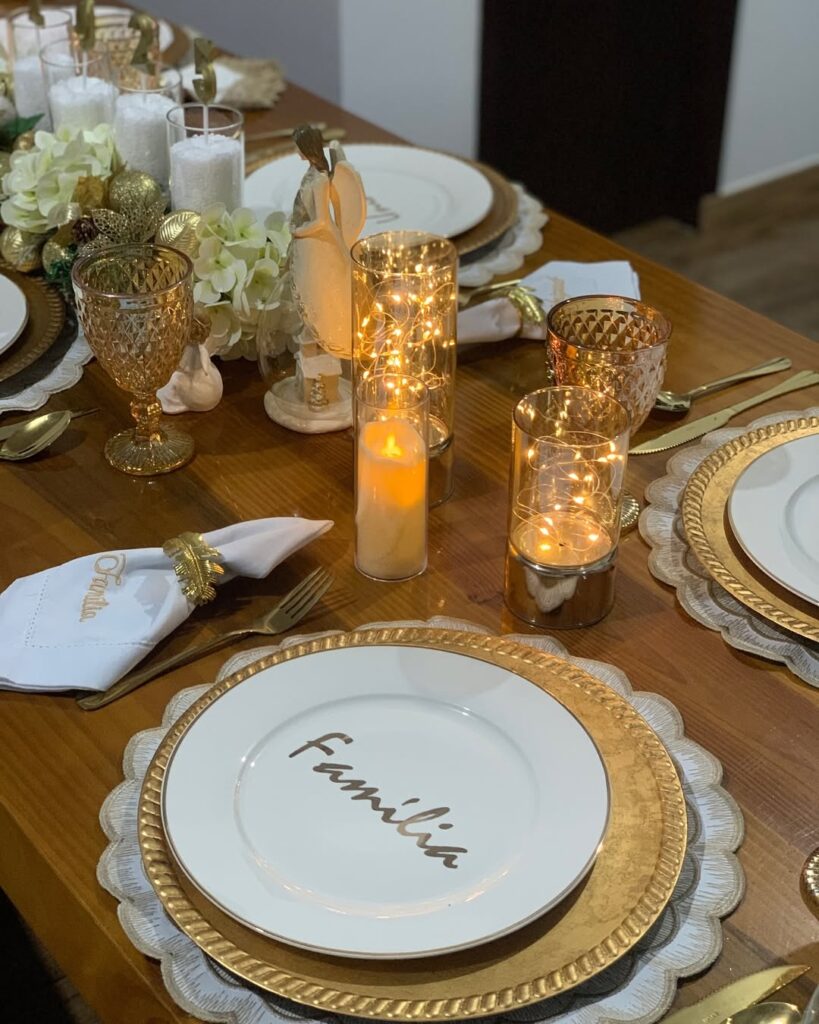 Mesa de madeira decorada com sousplat, prato dourado e outro branco por cima, velas e luzinhas