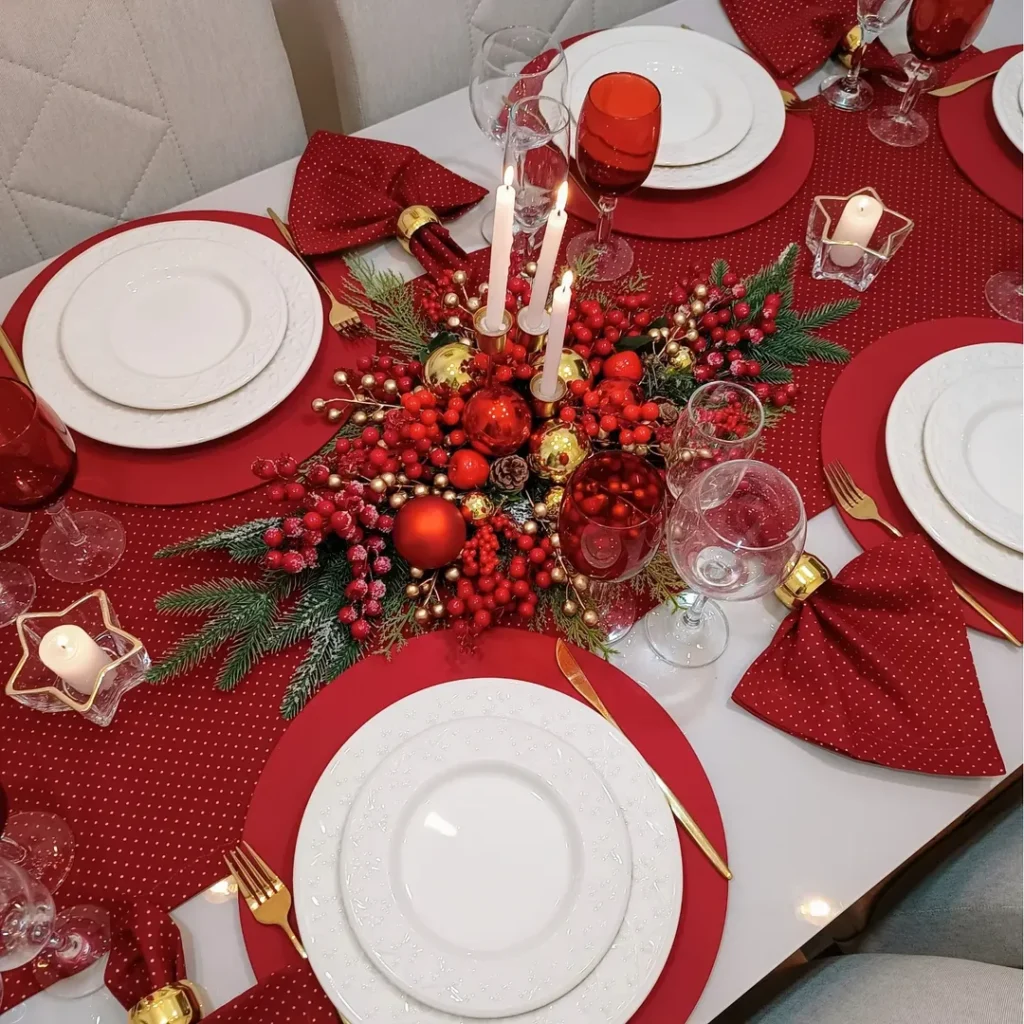 Mesa posta natalina com caminho de mesa e sousplats vermelhos, pratos brancos, talheres dourados e arranjo com velas, bolinhas e azevinhos