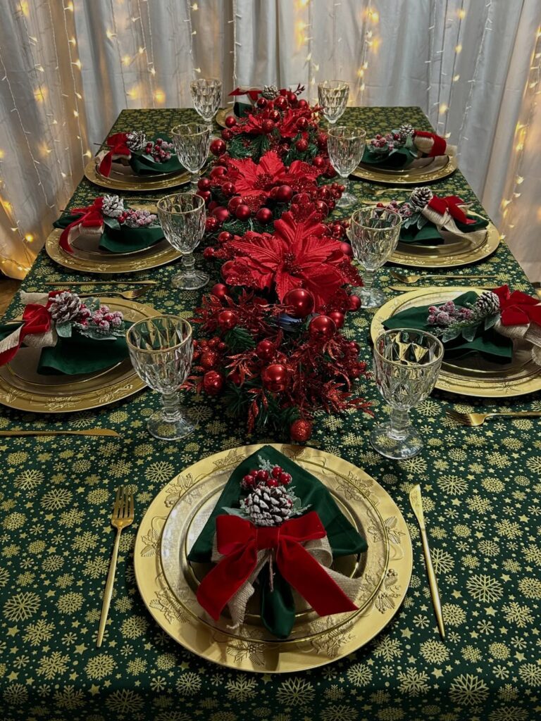 Mesa posta de Natal com toalha verde e desenhos dourados, sousplats e talhares dourados, guardanapos com pinhas decoradas e centro de mesa vermelho