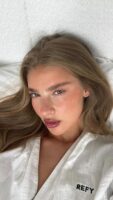 Mulher de pele clara, com roupão branco, cabelo claro e lash lifting