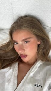 Mulher de pele clara, com roupão branco, cabelo claro e lash lifting