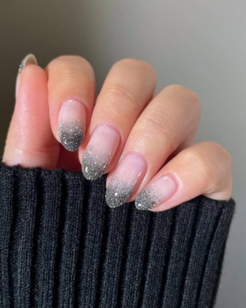 Mão de pele clara, com unha amendoada neutra com glitter prata e estilo ombré 