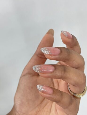 Mão de pele clara, com anel dourado e unha amendoada com ombré com glitter