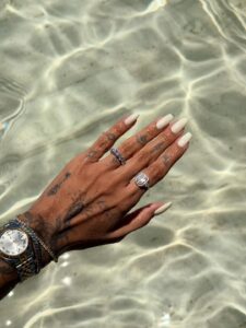 Mão de pele clara, com tatuagens, anéis e unha quadrada branca