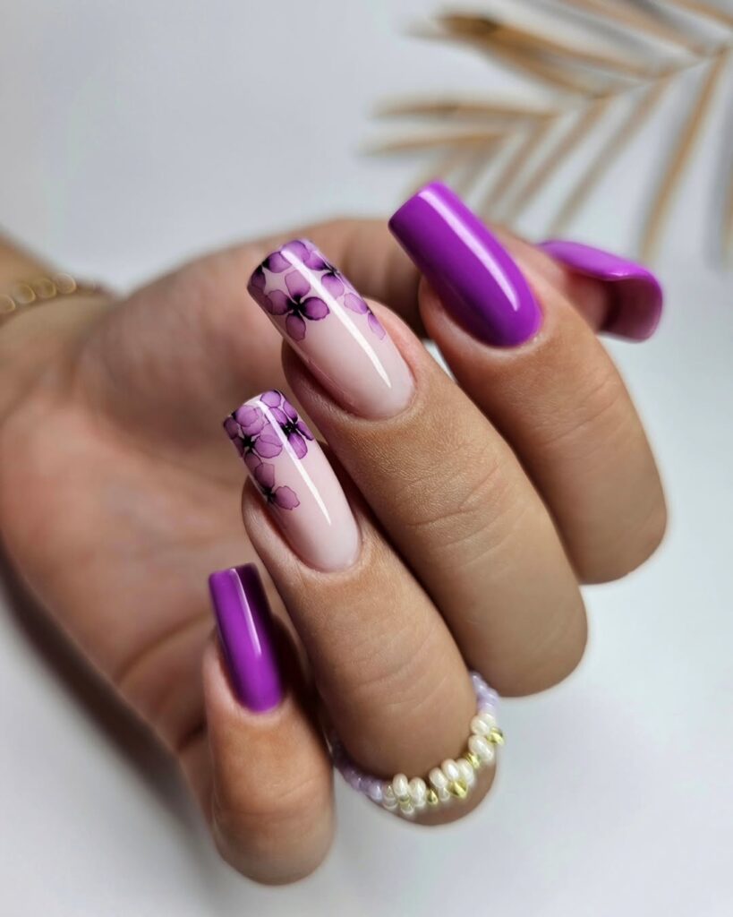 Mão de pele clara, com anel dourado e unha quadrada violeta com decoração de flor