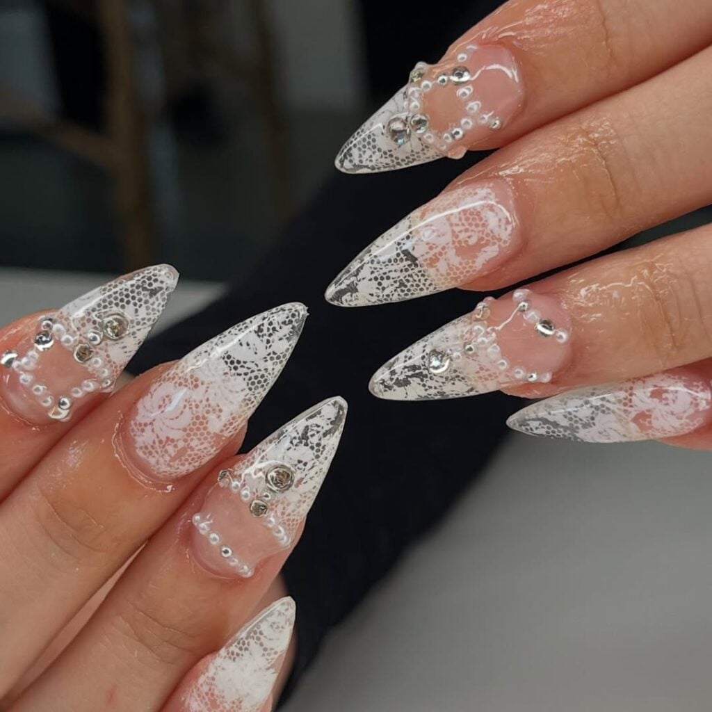 Mão de pele clara, com unha estilo stiletto e decoração com renda, pérolas e strass