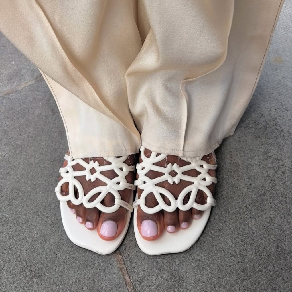 Pés de pele preta, com chinelo claro e unhas lilases